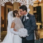 Elopement Wedding A bride and groom share a kiss under an ornate, white, wrought-iron arch. The bride wears a detailed lace dress and veil, holding a bouquet of white roses, while the groom is in a grey suit with a white rose boutonniere. The background is a grand building with lit steps and intricate decor. Elopements Inc